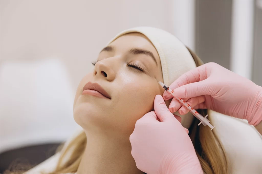 A woman receiving a cosmetic facial injection near her eye from a practitioner wearing pink gloves.